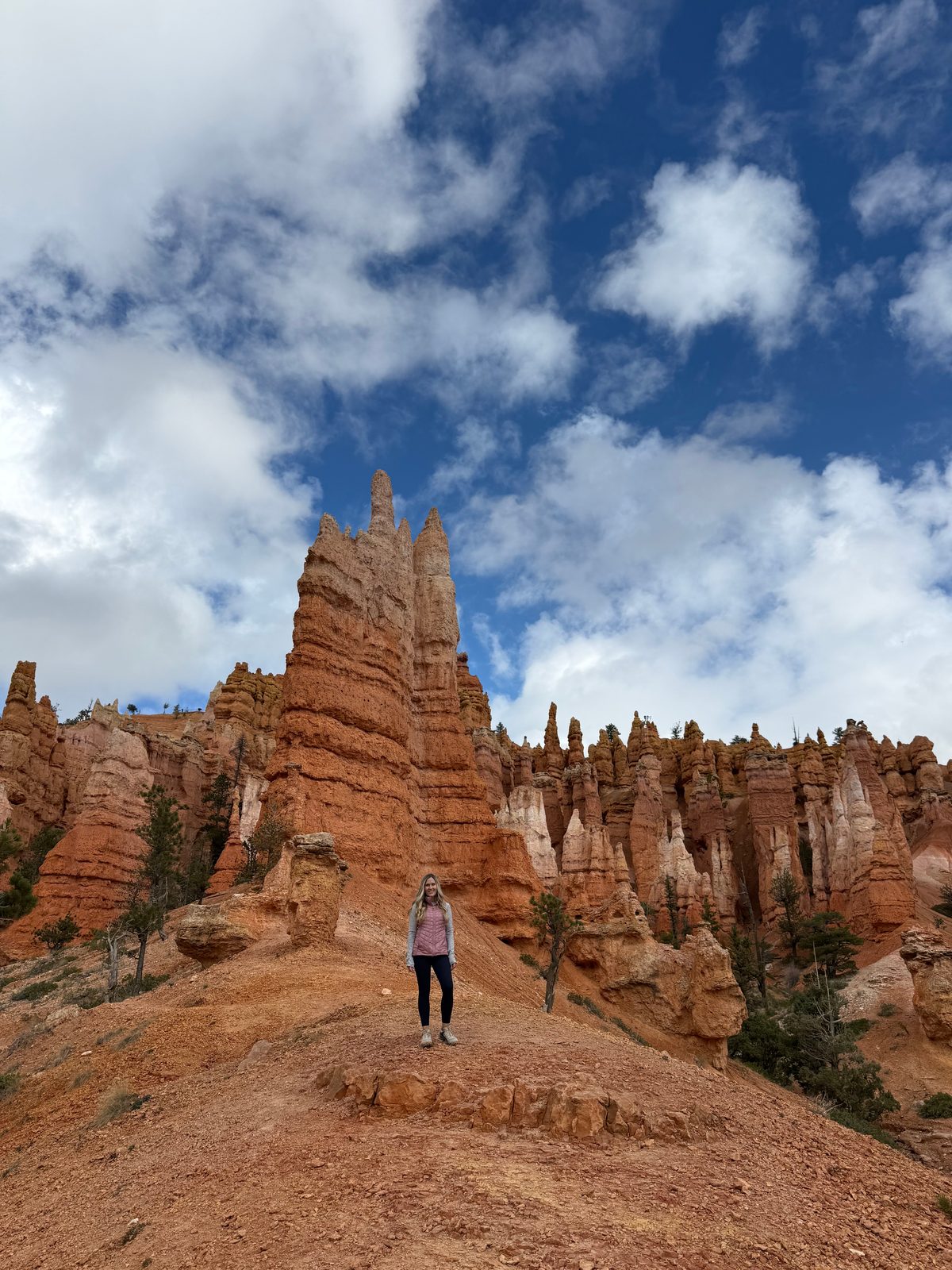 Trisha standing on an orange dirt ridge below a tall layered hoodoo spire, a row of smaller hoodoos behind her against a blue sky with white clouds