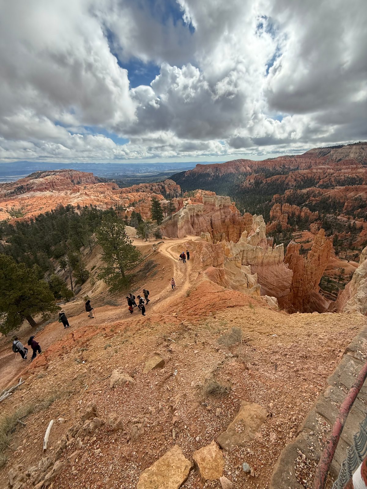Hikers climbing a switchbacking dirt trail up toward the rim near Sunrise Point, pale and orange hoodoos to the right and forested plateau and distant mesas under big clouds
