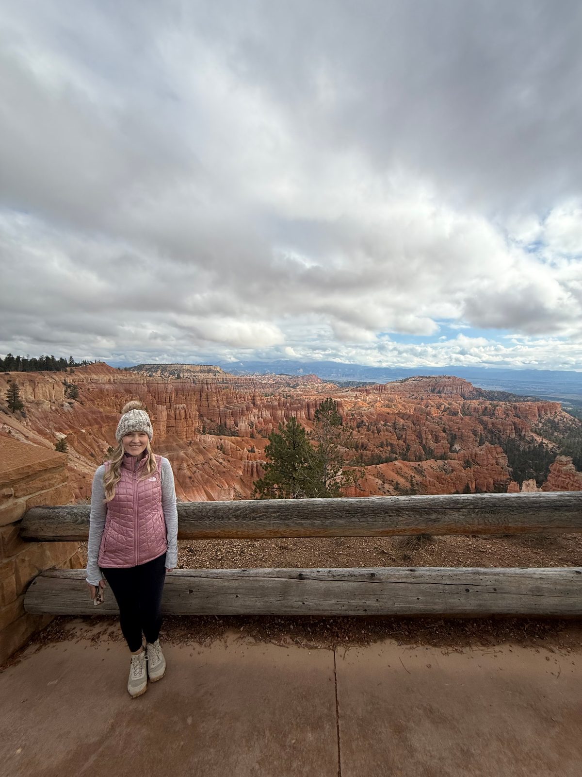 Trisha in a knit hat and pink puffer vest standing by a log railing at Sunset Point, the orange and white hoodoos of the amphitheater spread out behind her under heavy white clouds