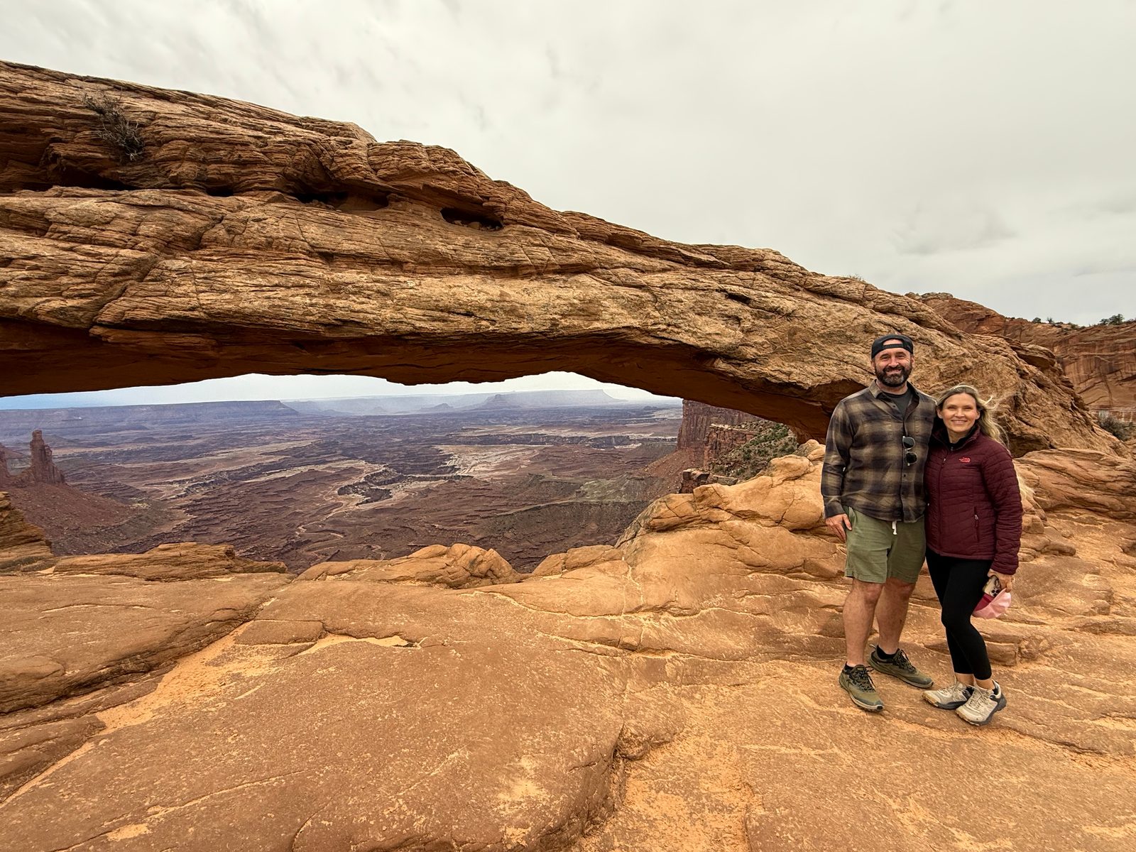 Tony and Trisha standing on slickrock beside Mesa Arch, a long low sandstone arch on the edge of a cliff framing a wide canyon landscape under a pale sky