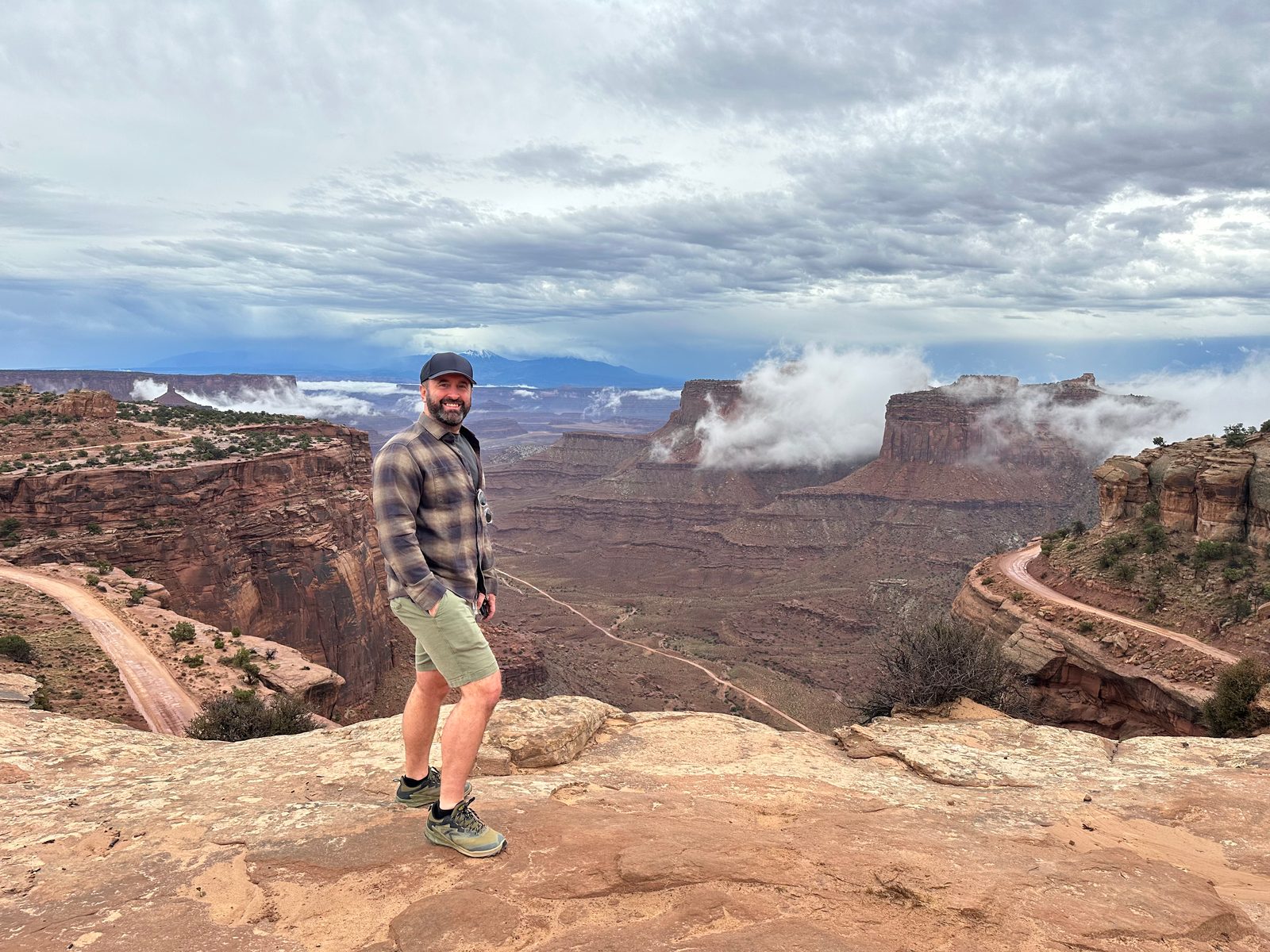 Tony standing on a sandstone ledge above a deep canyon, a dirt road winding down the cliff face below, mesas in the distance with low clouds caught against them
