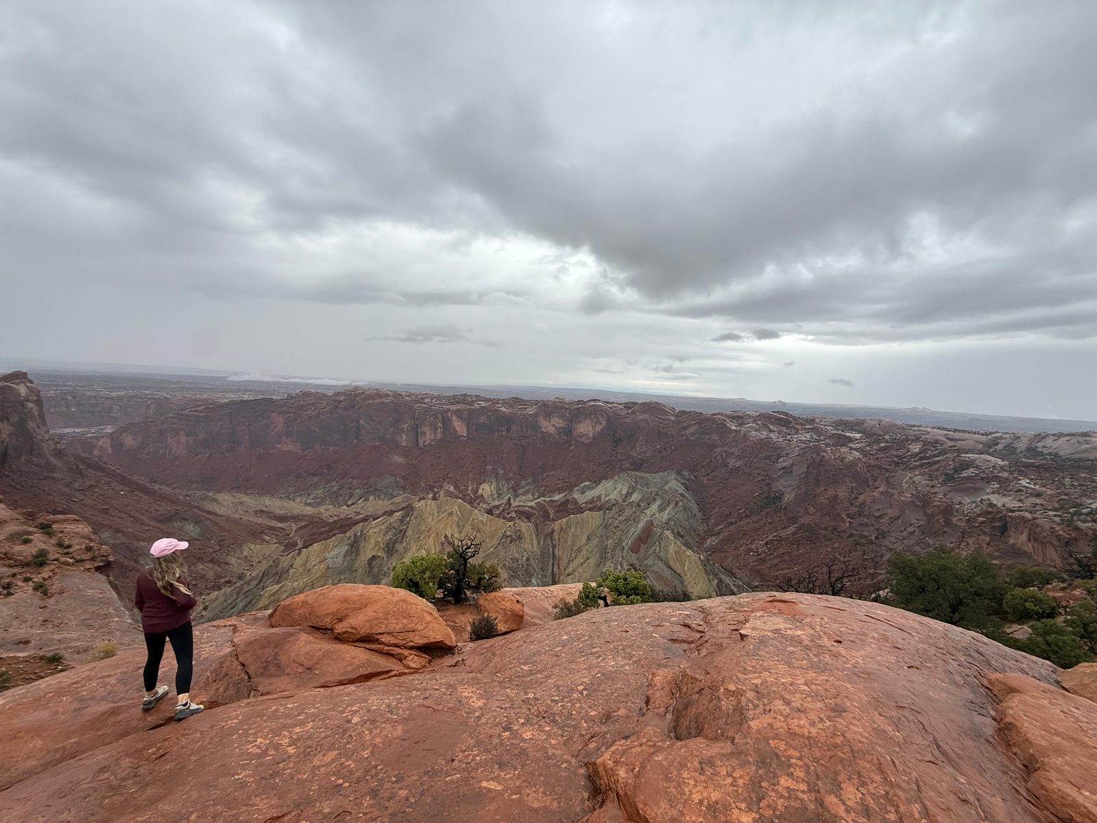 Trisha in a pink cap standing on red slickrock looking out over Upheaval Dome, a wide crater with gray-green rock heaped in its center and red cliffs around it under dark clouds