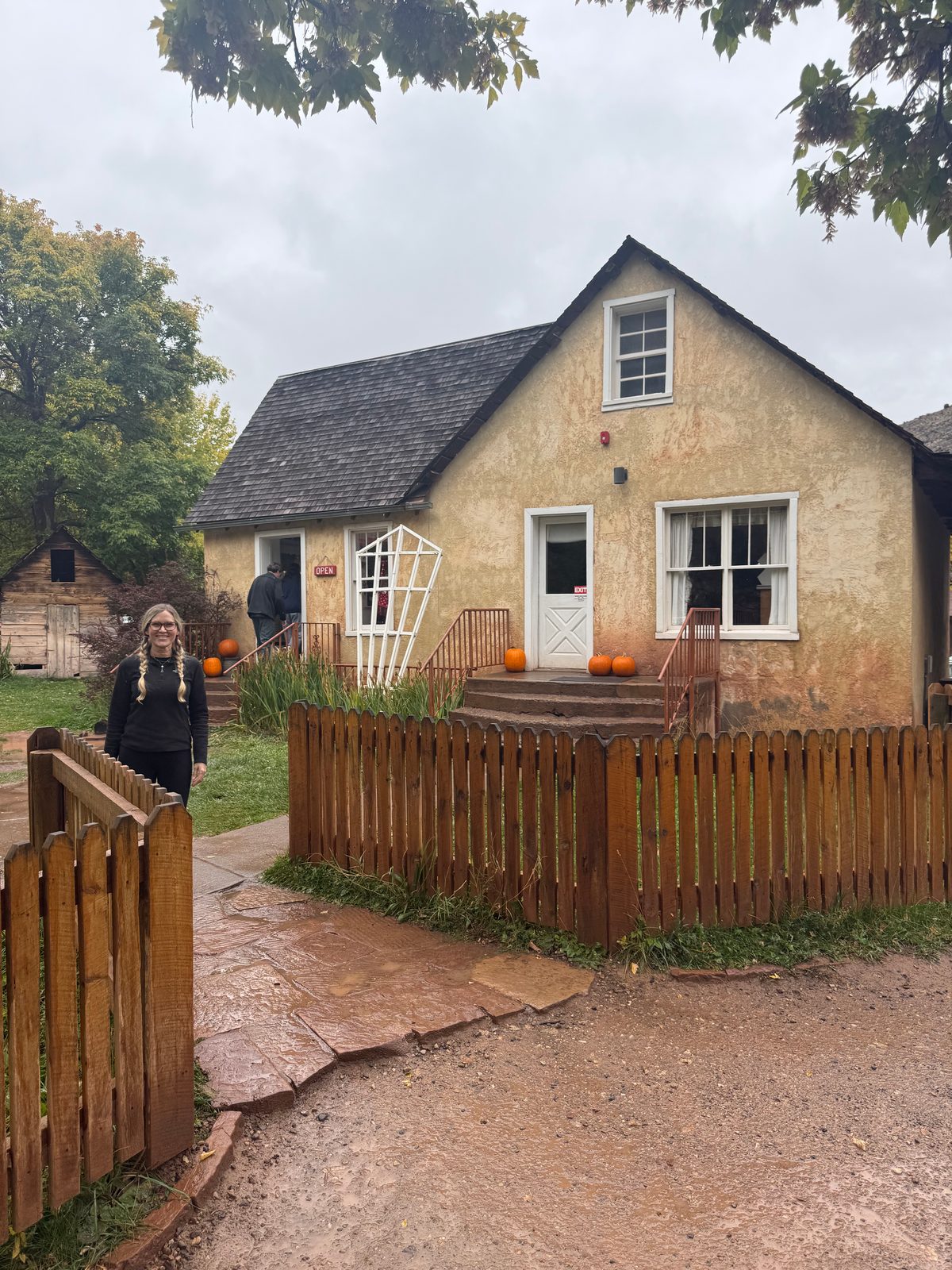 Trisha at the gate of a wooden picket fence in front of the Gifford House, a cream stucco farmhouse with pumpkins on the steps and an open sign, wet flagstones and trees around it