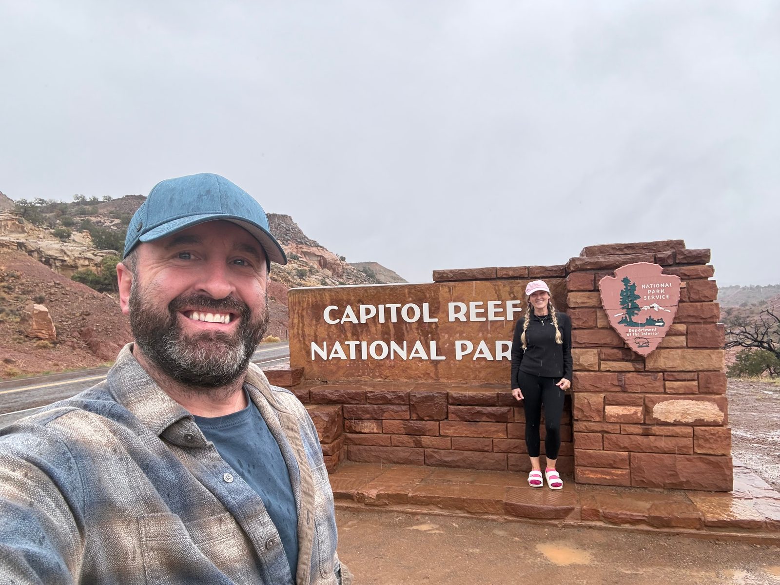 Selfie of Tony in a rain-spotted cap and flannel shirt, Trisha behind him at the red stone Capitol Reef National Park entrance sign, wet road and gray sky