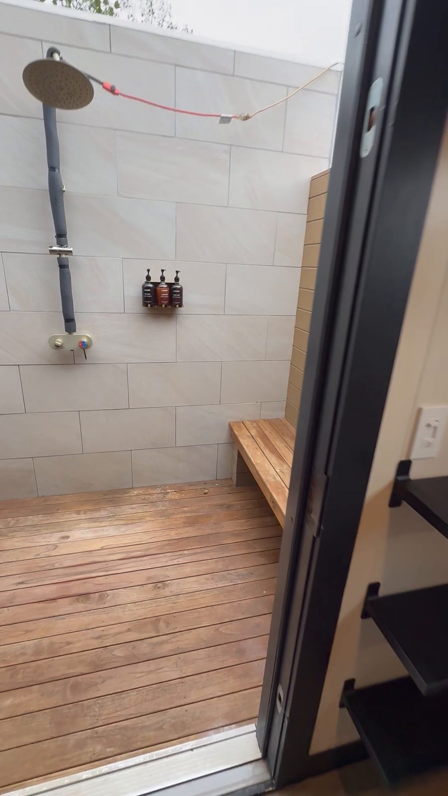 An open-air shower beside a cabin at Ofland Escalante: a rain showerhead on a pale tiled wall, a wooden bench, a slatted wood floor and open sky above
