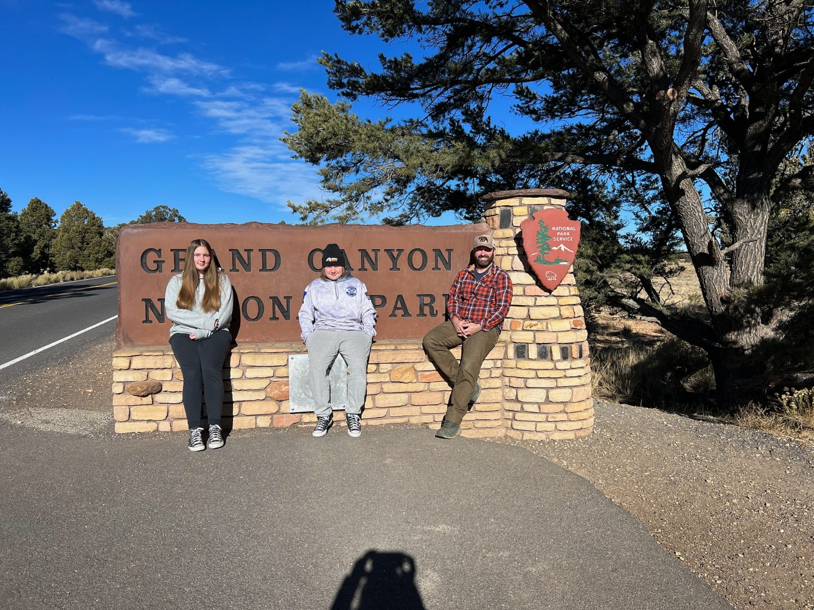 Lily, Landon and Tony sitting along the stone base of the Grand Canyon National Park entrance sign, with the National Park Service arrowhead beside them