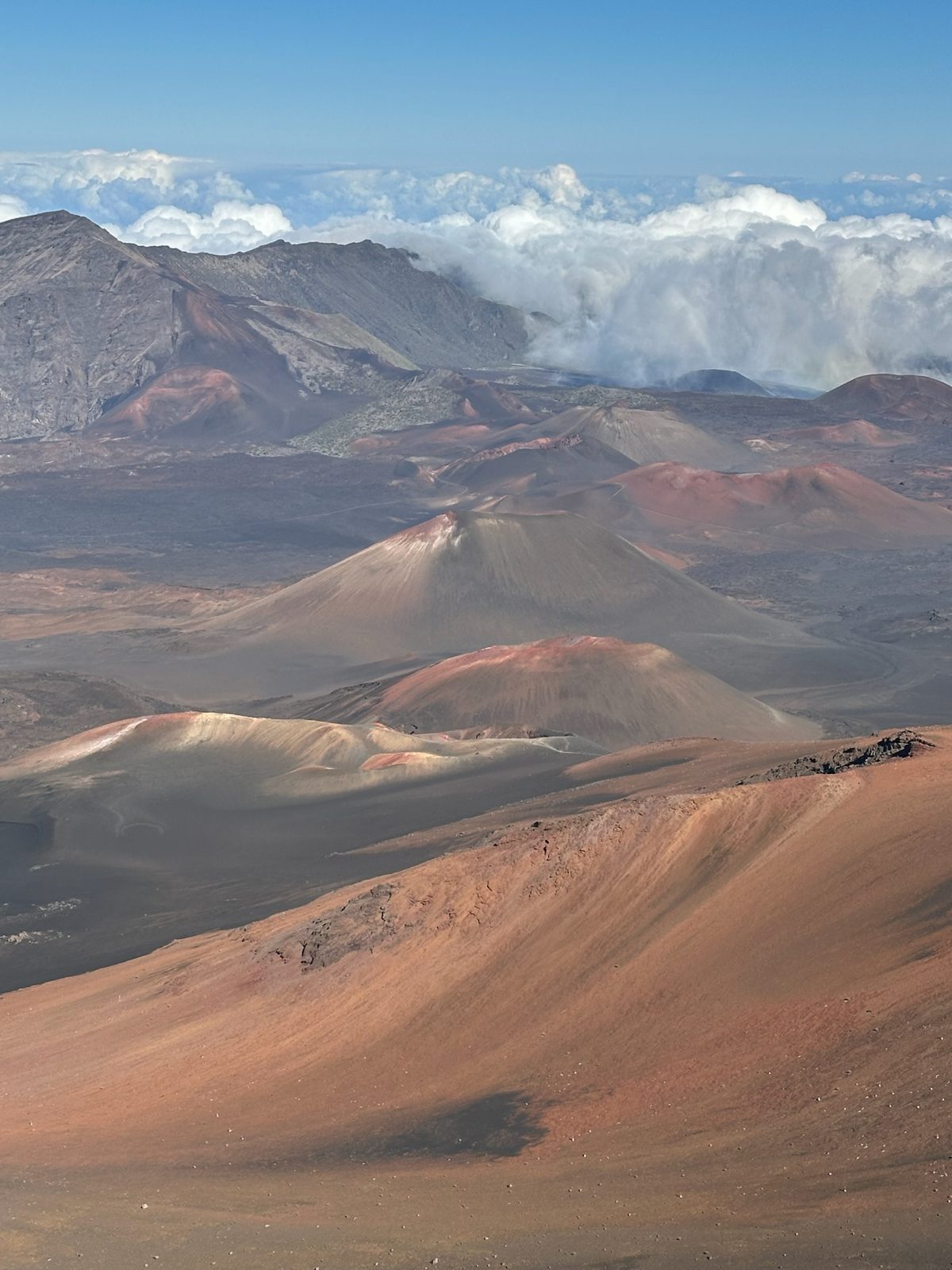 The floor of Haleakala crater, red and grey cinder cones running into the distance with cloud pouring over the far rim