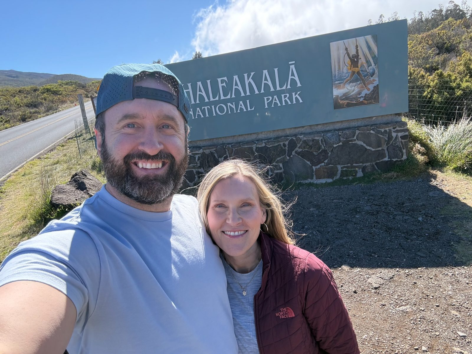 Tony and Trisha at the Haleakala National Park entrance sign, which carries a painting of the demigod Maui lassoing the sun