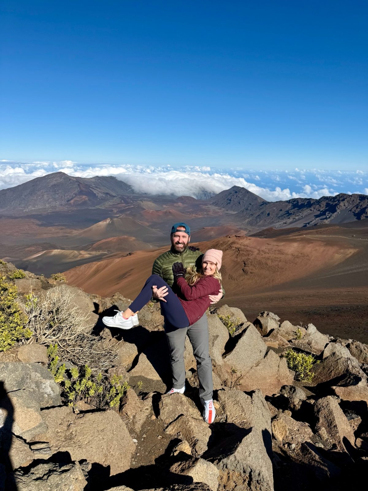 Tony holding Trisha at the Haleakala summit, the cinder cone floor of the crater behind them and a solid deck of cloud below the far rim