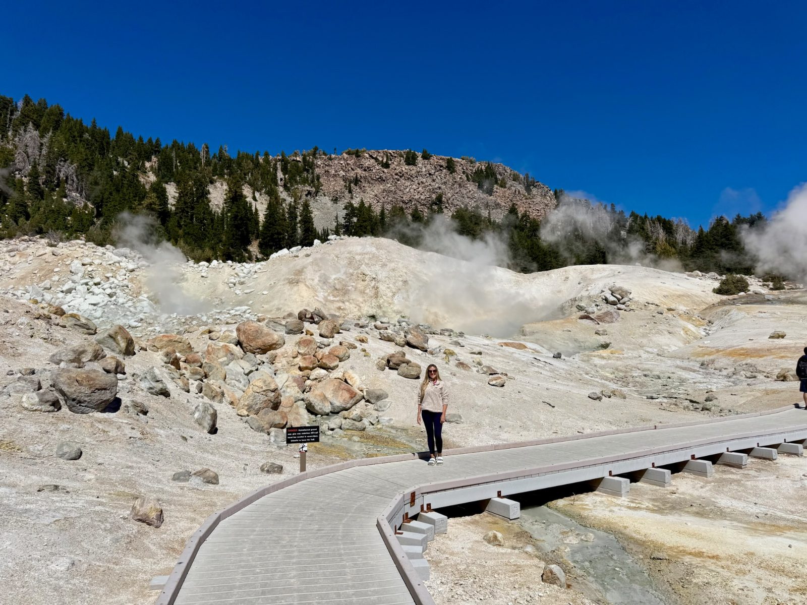 Trisha standing on the boardwalk through Bumpass Hell, steam venting from the white ground on both sides, a hydrothermal danger sign beside the walkway