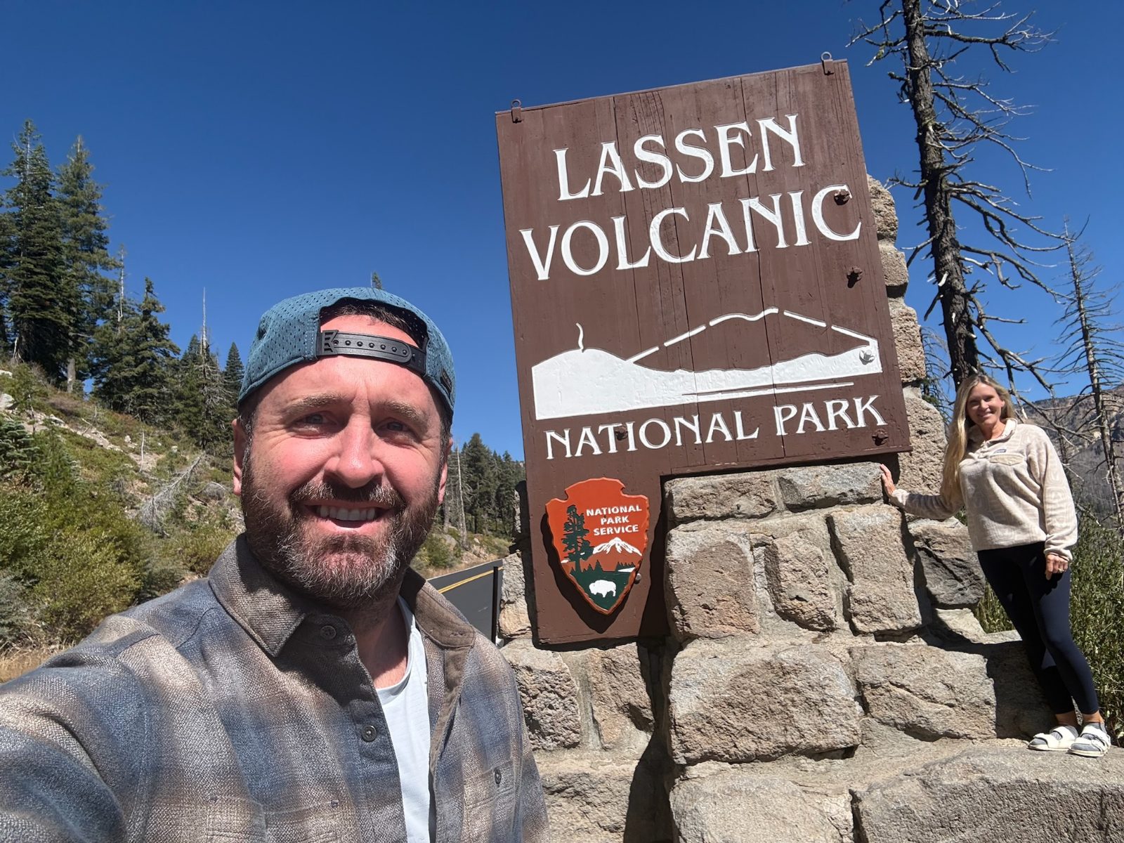 Tony and Trisha at the wooden Lassen Volcanic National Park entrance sign, pines and blue sky behind