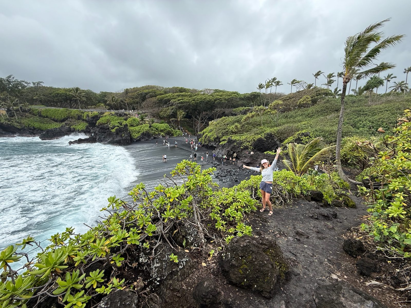 Trisha standing with her arms out on the lava cliff above the black sand beach at Waianapanapa, surf breaking white against the dark sand