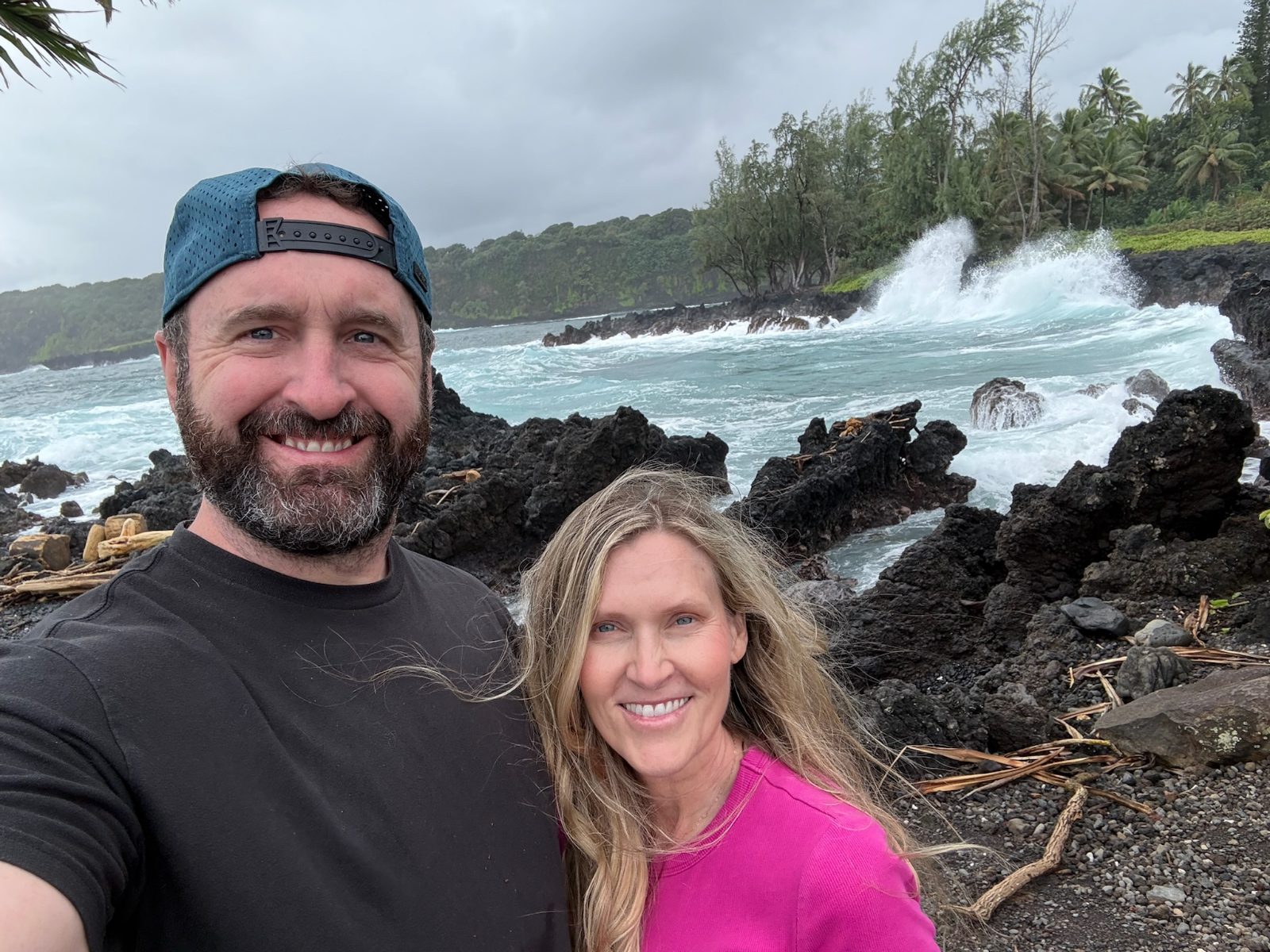 Tony and Trisha on the black lava shoreline at Keanae, heavy surf exploding on the rocks behind them under a grey sky