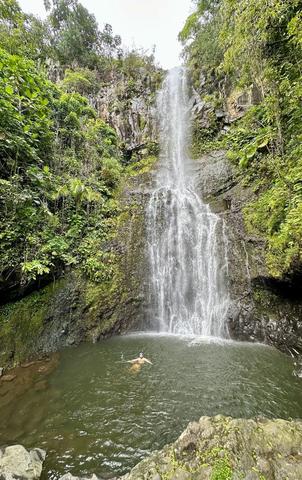 Tony swimming in the pool at the base of a tall waterfall on Maui, rainforest and rock on both sides