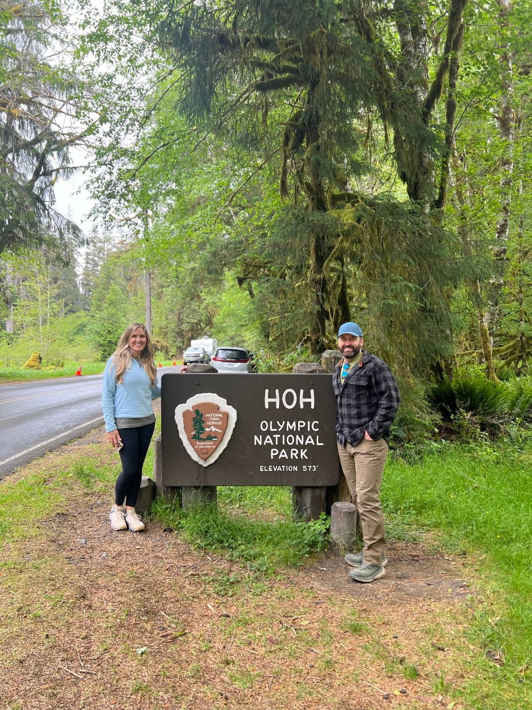 Tony and Trisha standing either side of the brown Hoh Olympic National Park entrance sign, moss-hung trees behind