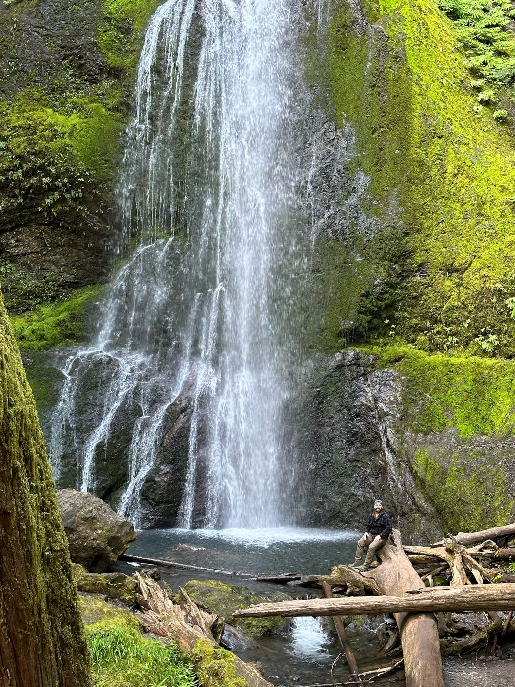 Tony sitting on driftwood at the base of Marymere Falls, water dropping down a tall moss-covered rock face