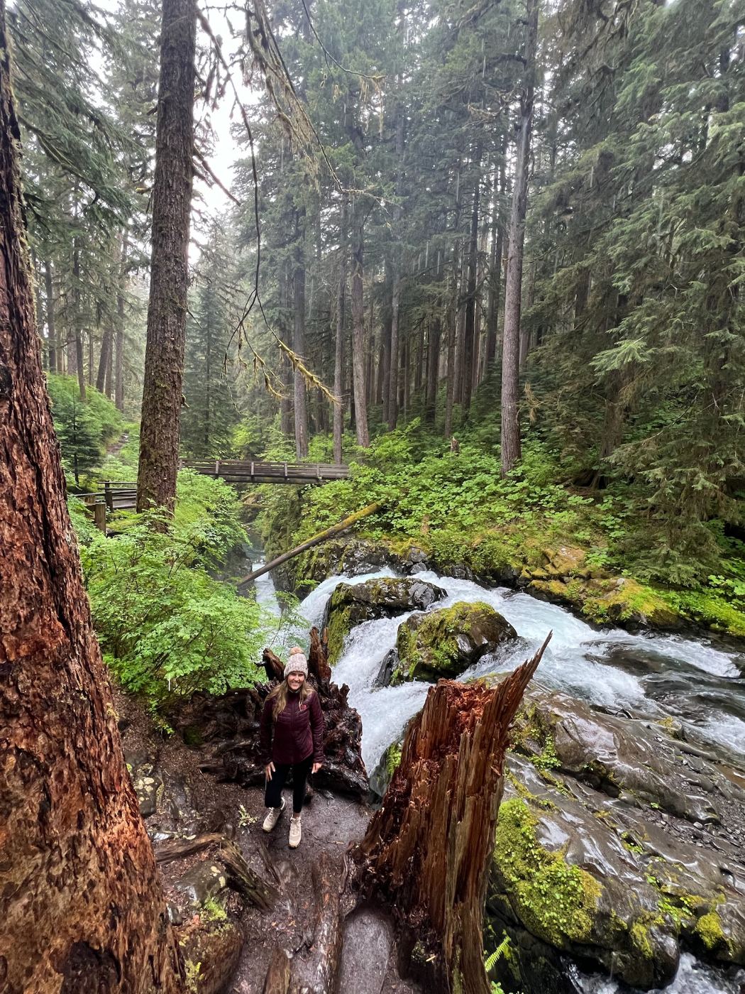 Trisha in a beanie and jacket beside the Sol Duc river, a wooden footbridge crossing the gorge upstream