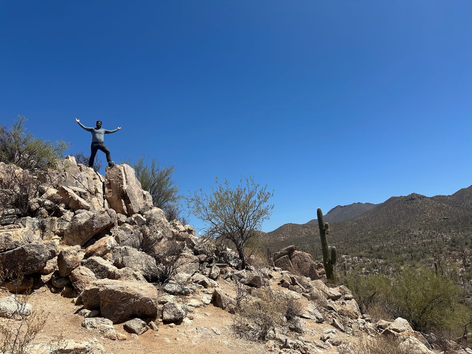 Tony standing arms out on a granite outcrop against a deep blue sky, saguaro and desert mountains behind