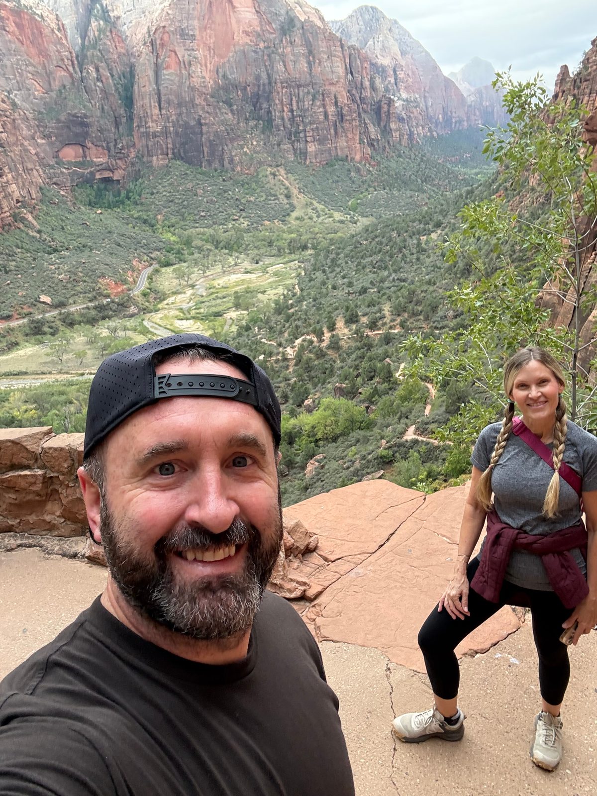 Selfie of Tony in a backwards cap with Trisha behind him on a sandstone ledge, Zion Canyon spread out below them with the river, the road and tall red cliffs