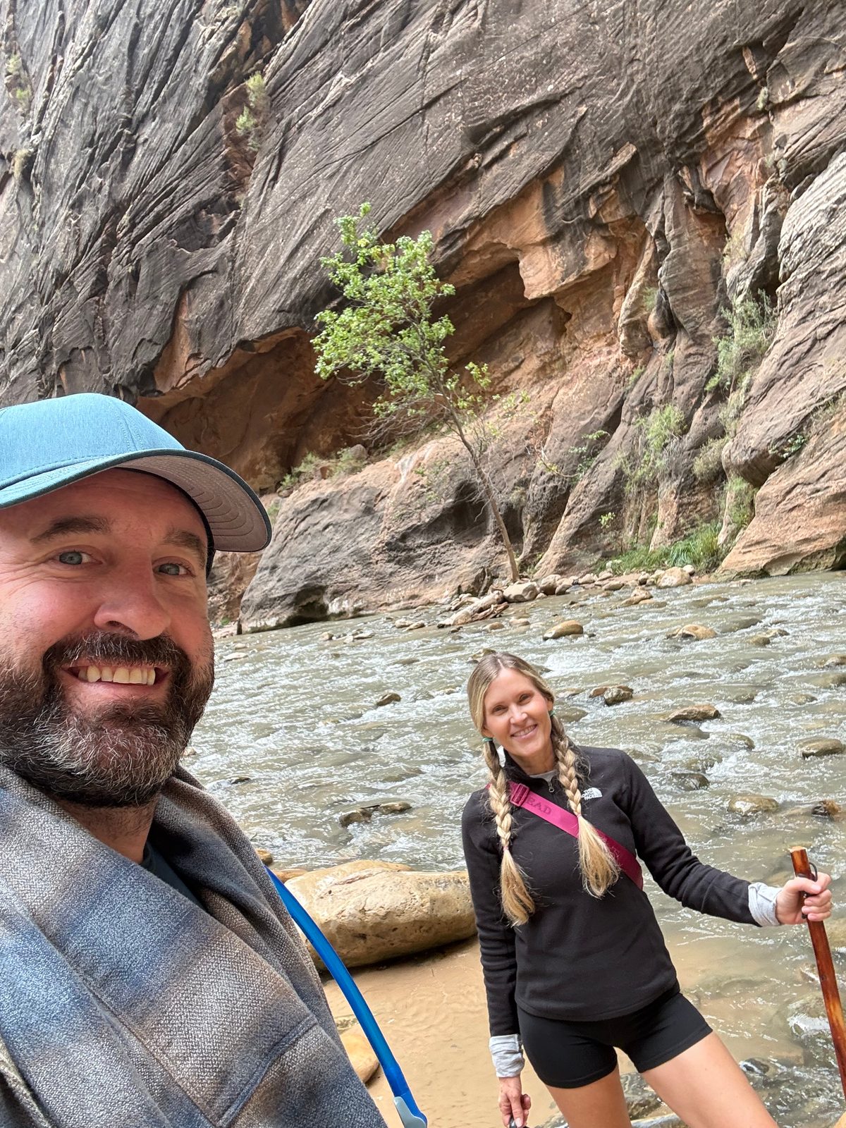 Selfie of Tony in a cap and flannel shirt with Trisha behind him holding a walking stick, both smiling, the river and a sheer canyon wall with a small tree behind them
