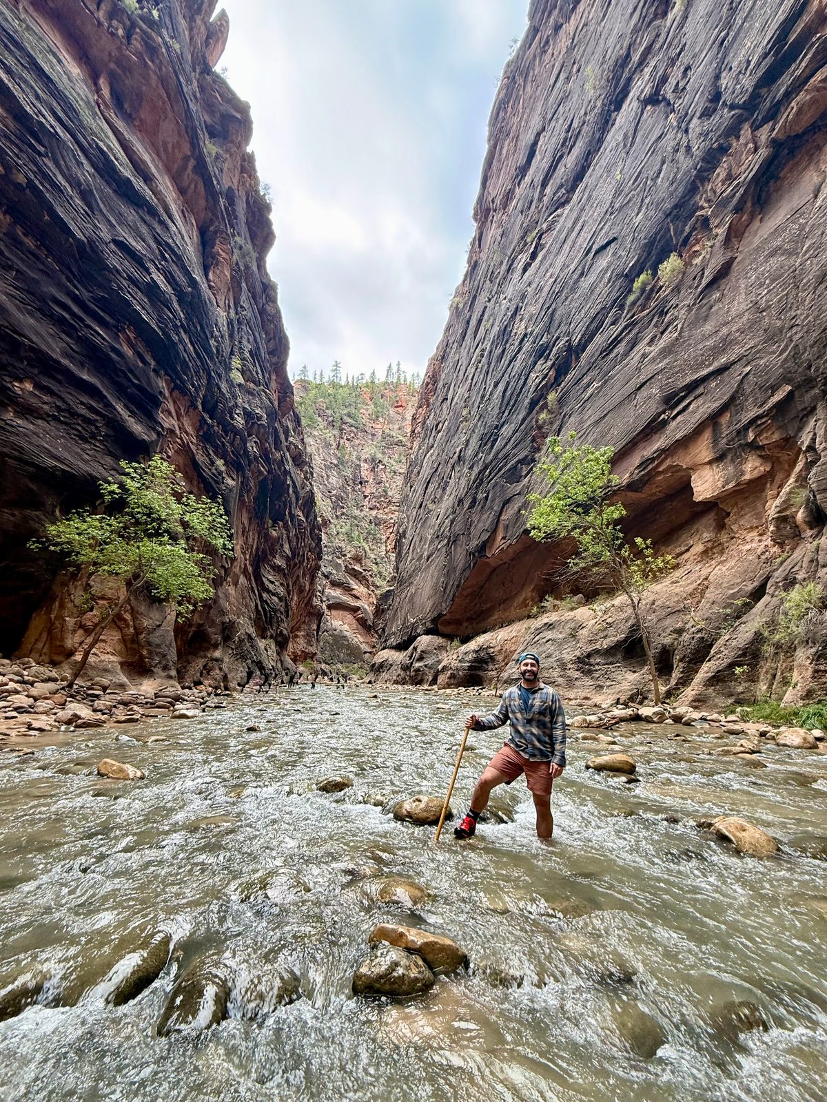 Tony standing in the Virgin River in the Zion Narrows, holding a wooden walking stick, the water around his shins and sheer dark canyon walls rising on both sides
