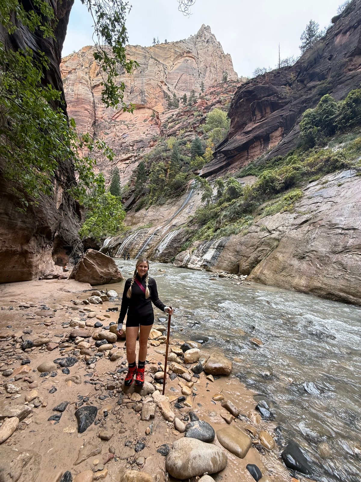 Trisha on a gravel bar beside the river in the Narrows, wearing red neoprene socks and holding a wooden walking stick, a tall streaked sandstone wall behind her