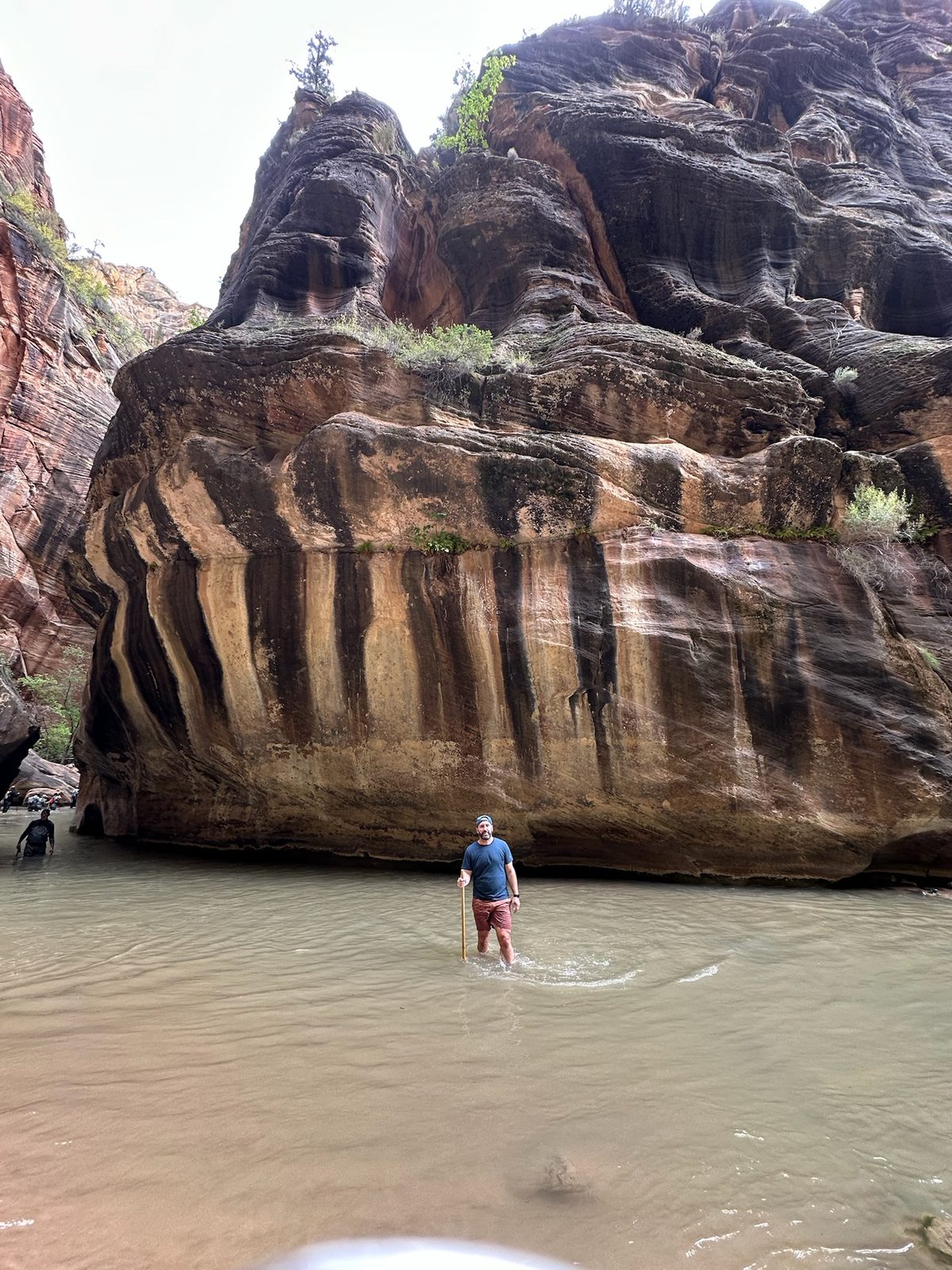 Tony standing knee-deep in calm brown water in front of a massive sandstone wall striped vertically with dark streaks, a hiker wading in the distance