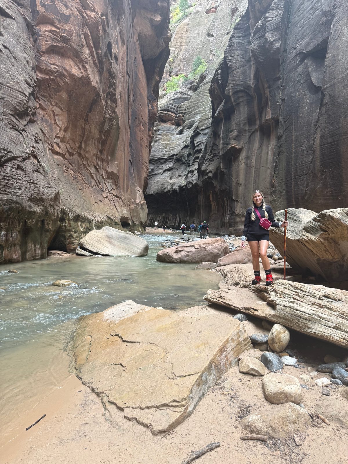 Trisha standing on a sandstone ledge beside the river where the Narrows tightens, smooth dark walls rising high on both sides and a line of hikers in the water behind her