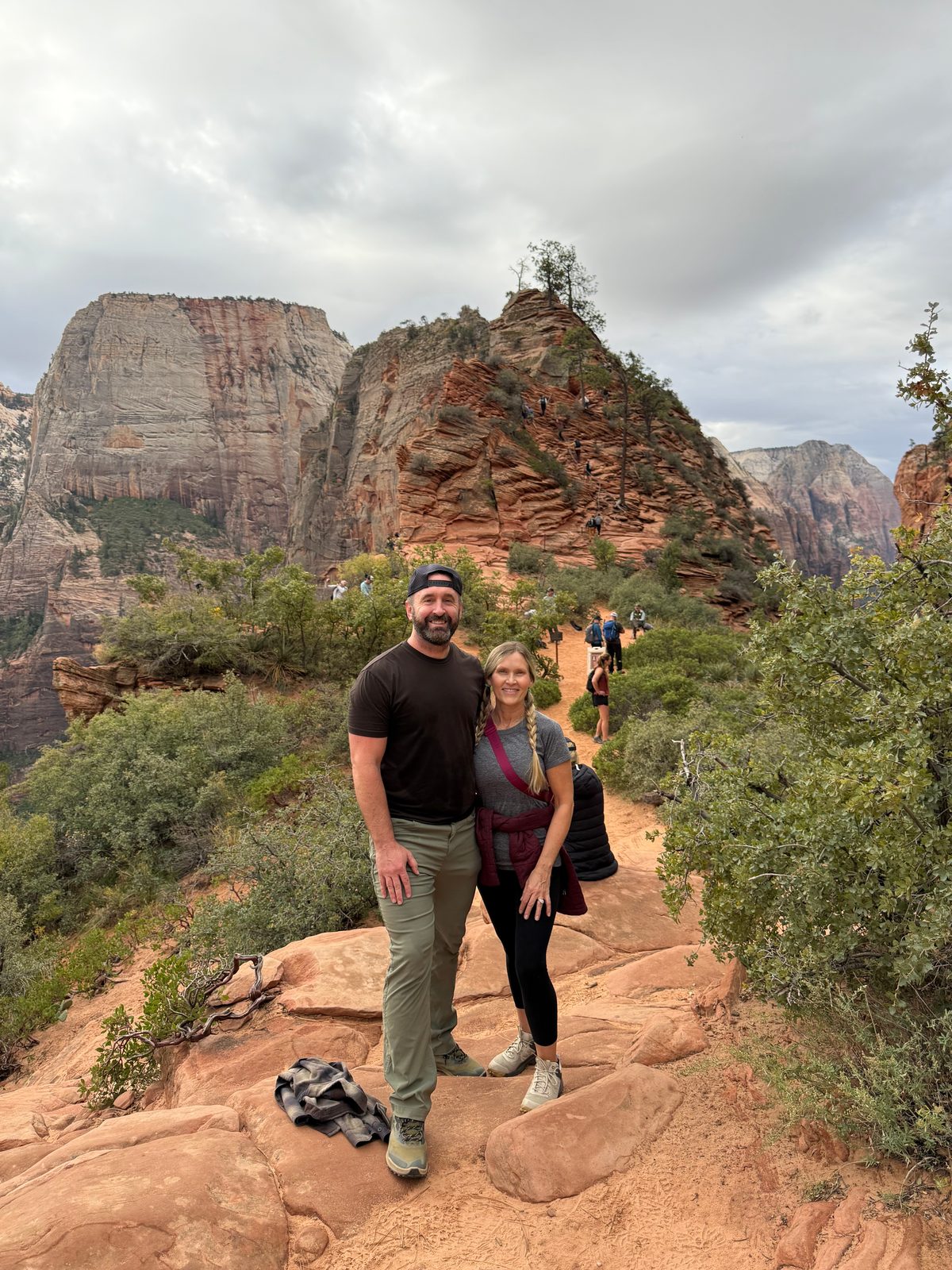 Tony and Trisha standing together on red rock at Scout Lookout, the narrow fin of Angels Landing rising behind them with hikers climbing it, a pale sandstone cliff to the left under a gray sky