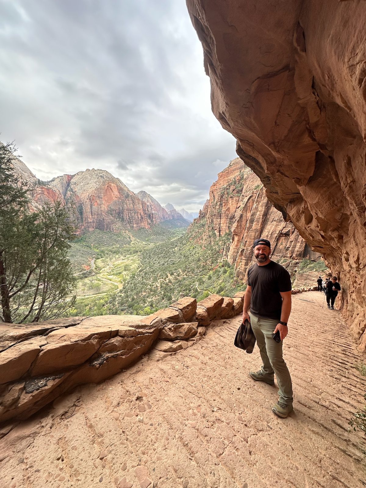 Tony on a wide trail cut into the side of a red cliff under an overhang, the green floor of Zion Canyon and its peaks far below