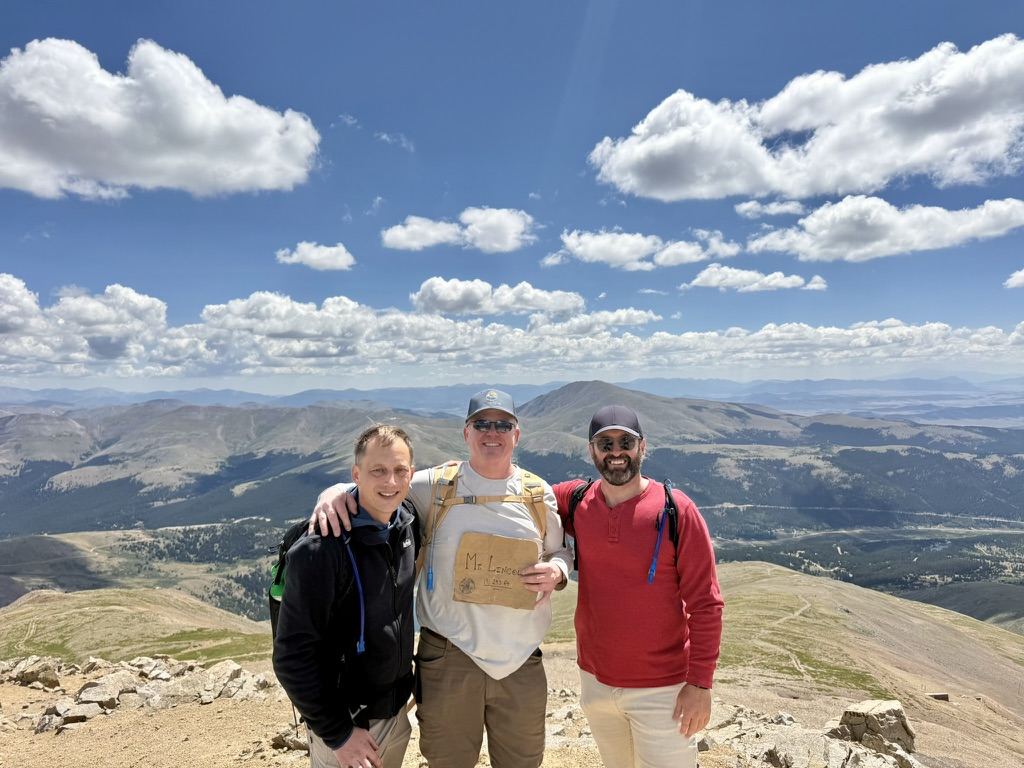 Three men on the summit of Mount Lincoln with a cardboard sign, mountains stretching to the horizon