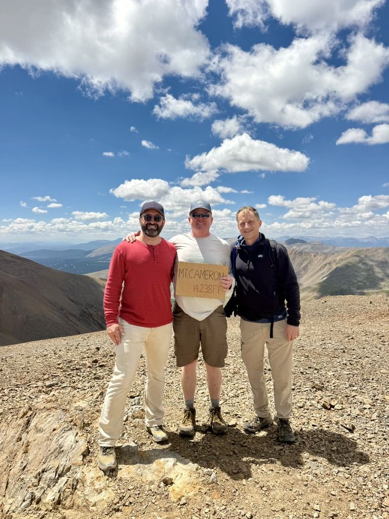 Three men on the summit of Mount Cameron holding a cardboard sign reading 14,238 feet
