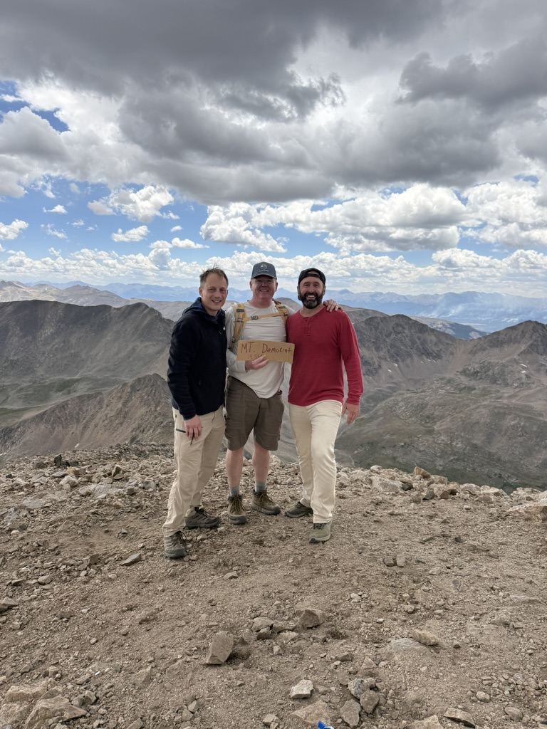 Three men on the summit of Mount Democrat holding a cardboard sign, clouds gathering behind them