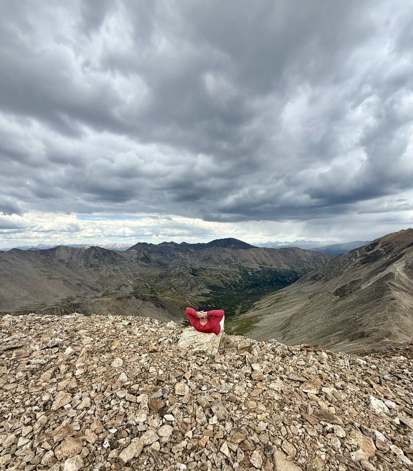 A hiker lying on the summit rocks looking out at a wide valley under heavy storm clouds