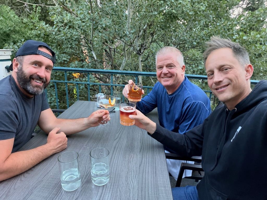 Three men toasting drinks at an outdoor table under aspen trees after the hike